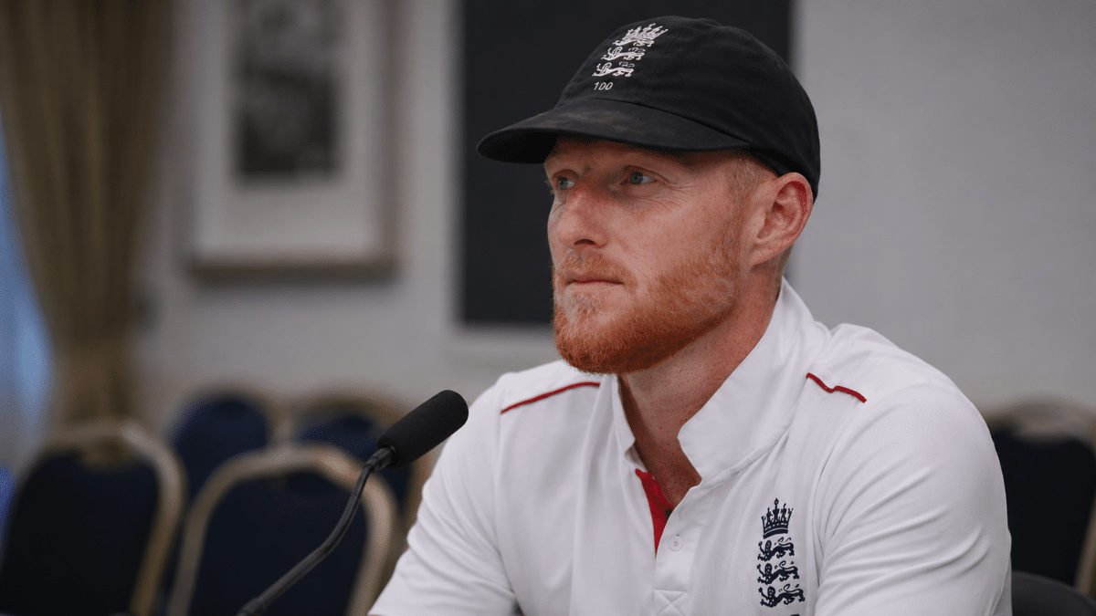 England cricketer Ben Stokes speaks during a press conference in a side-angle close-up photograph with a microphone and blurred media room background.