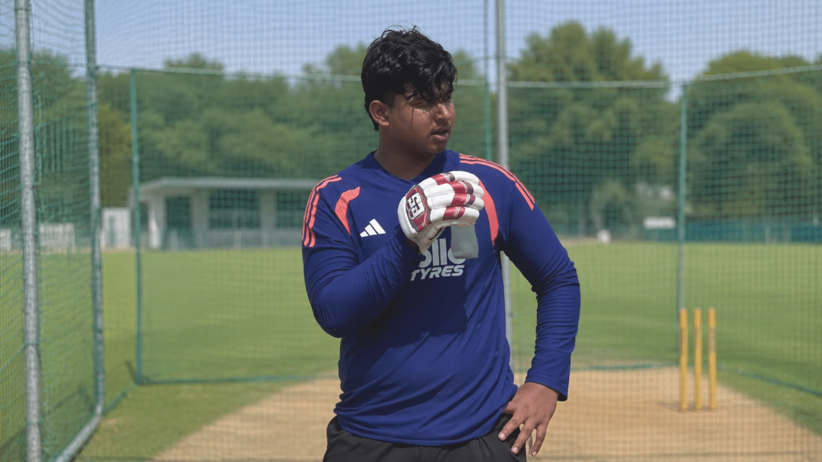 Vaibhav Sooryavanshi wearing a blue training jersey and Batting gloves standing inside outdoor cricket practice nets,