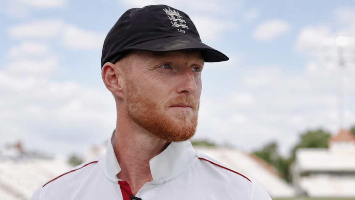 Close-up portrait of a professional cricketer wearing a black cap and white cricket jersey, looking to the side at an outdoor stadium with a softly blurred background and cloudy sky.