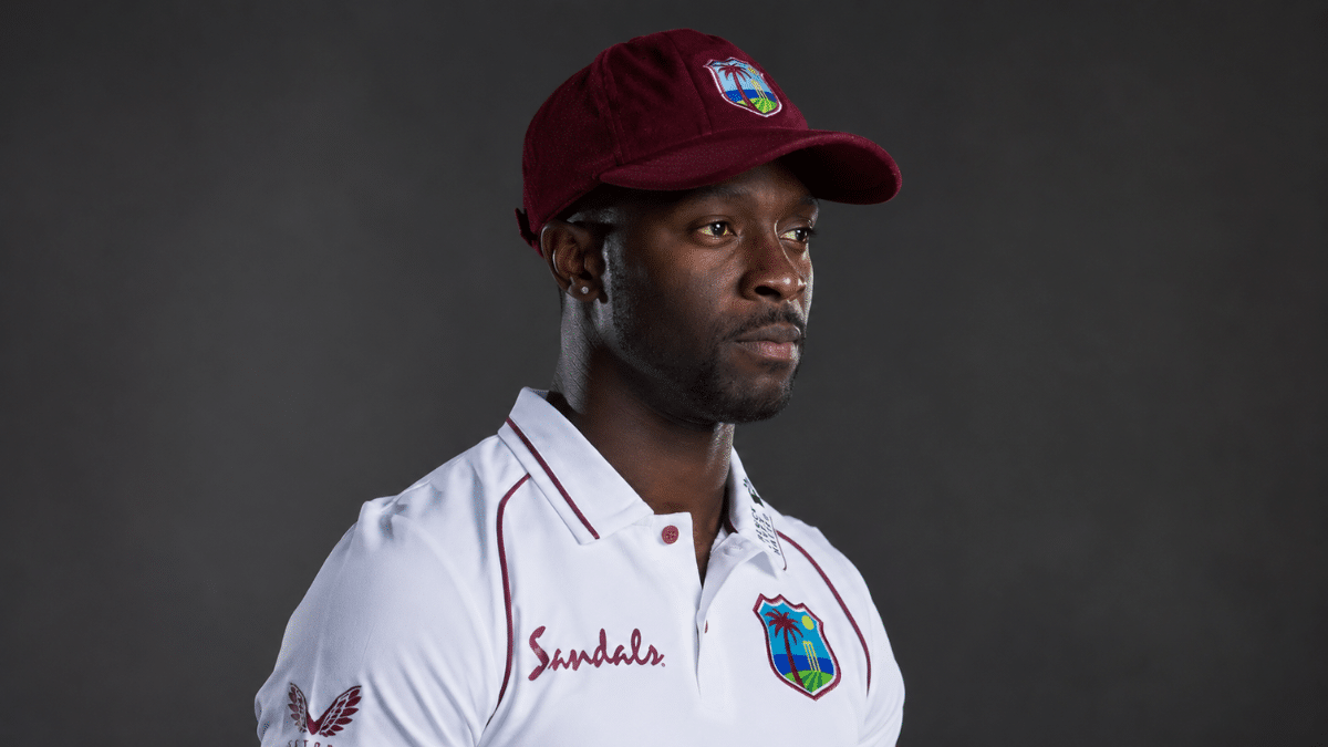 Kemar Roach Side-profile portrait of a professional cricketer wearing a maroon cricket cap and white Test cricket jersey, looking into the distance against a softly lit dark studio background.