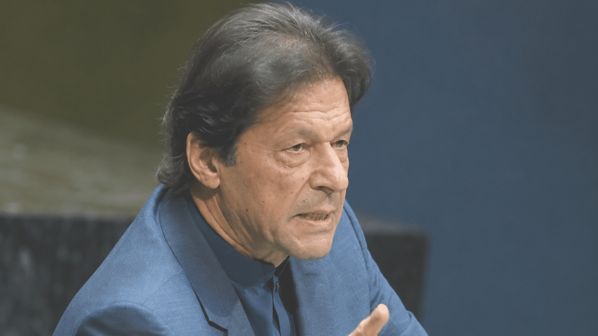 Imran Khan speaking at a podium, pointing forward with a serious expression, wearing a blue suit against a dark blue background.