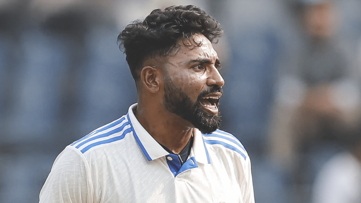 Indian cricketer shouting intensely during a match, wearing a white jersey with blue stripes, captured in side profile against a blurred stadium background.