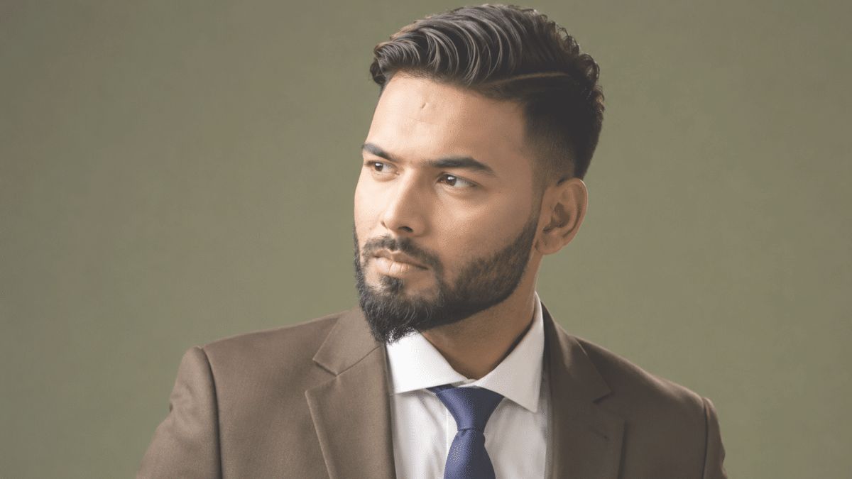 Young bearded man in a brown suit, white shirt, and navy tie, looking toward the left against an olive-green background.