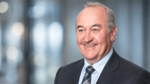Peter Roach in a dark suit and tie, smiling and looking to the side against a softly blurred blue background.