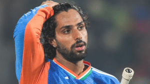 Tilak Varma holding a cricket bat and looking to the side in an India cricket jersey during a match.