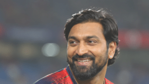 Kunal Pandya smiling in a navy-blue and red cricket jersey, shown at a right-facing angle with a blurred stadium background.