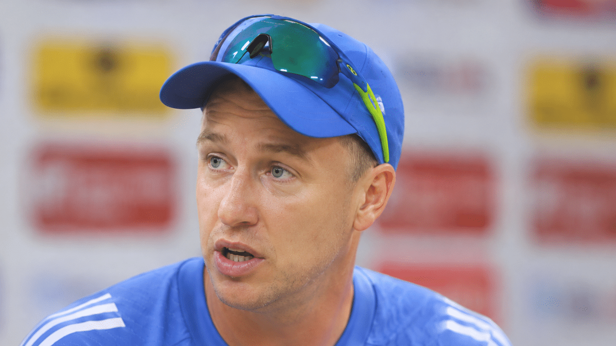 Morne Morkel speaking during a press conference, wearing a blue cricket training jersey and cap, with a blurred sponsor backdrop behind him.