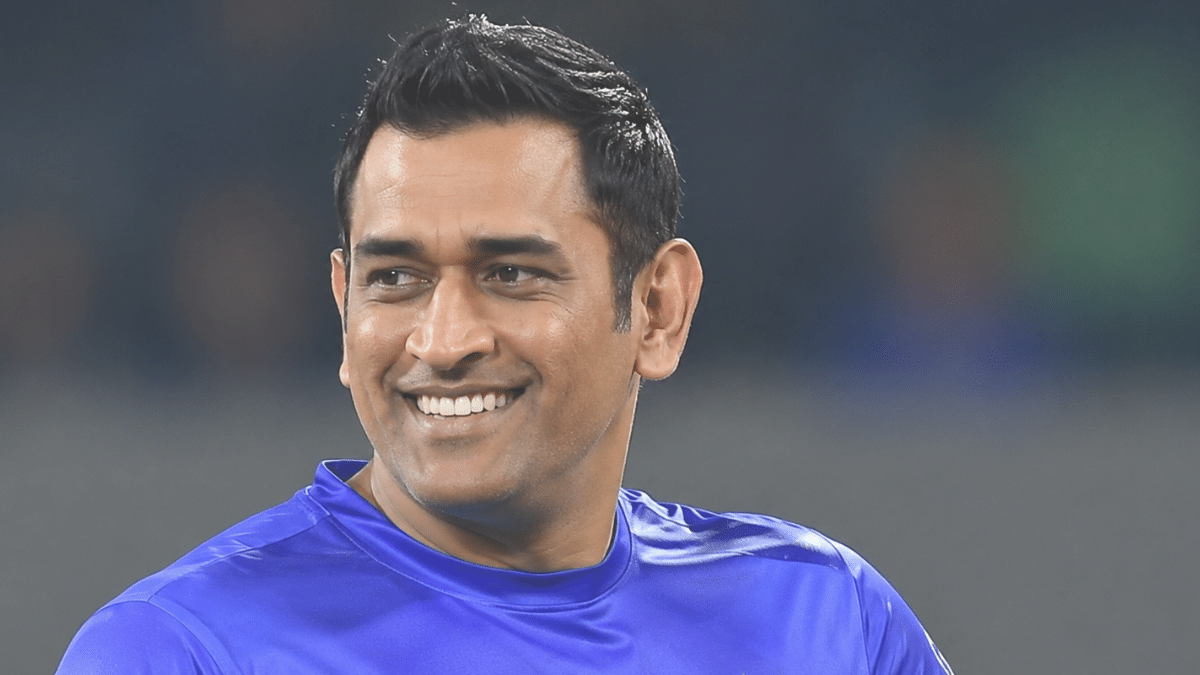MS Dhoni smiling and looking to the left in a blue cricket jersey during a stadium setting.