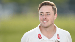 Ollie Robinson smiling and looking to the side in a white cricket shirt against a blurred green cricket field.