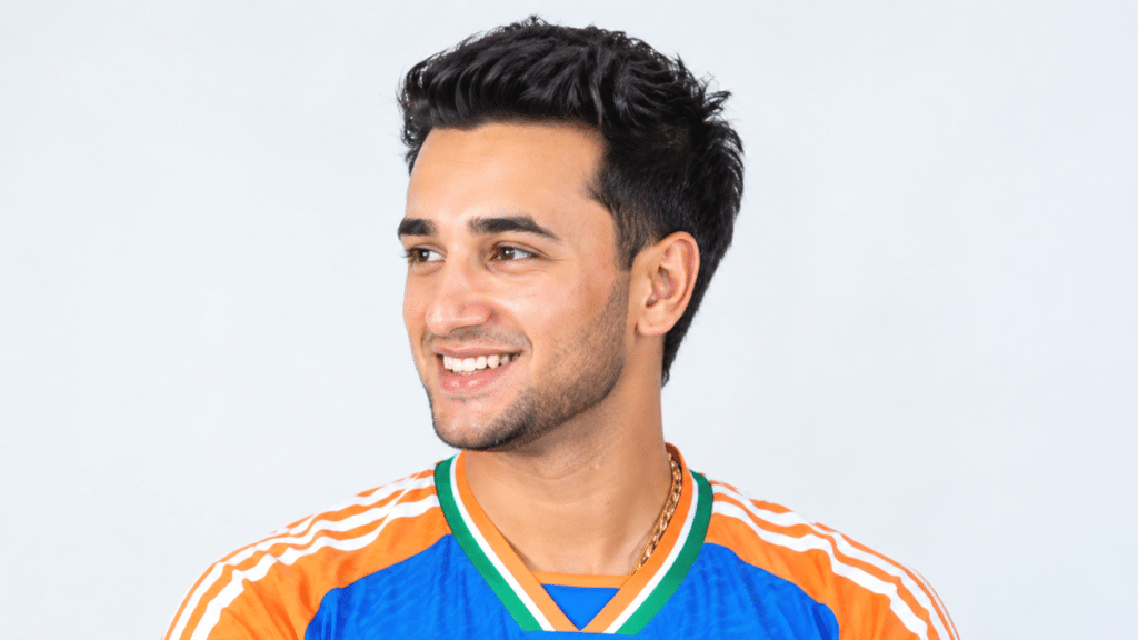 Abhishek Sharma smiling in an India cricket jersey, looking to the left in a three-quarter profile against a light background.