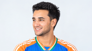 Abhishek Sharma smiling in an India cricket jersey, looking to the left in a three-quarter profile against a light background.