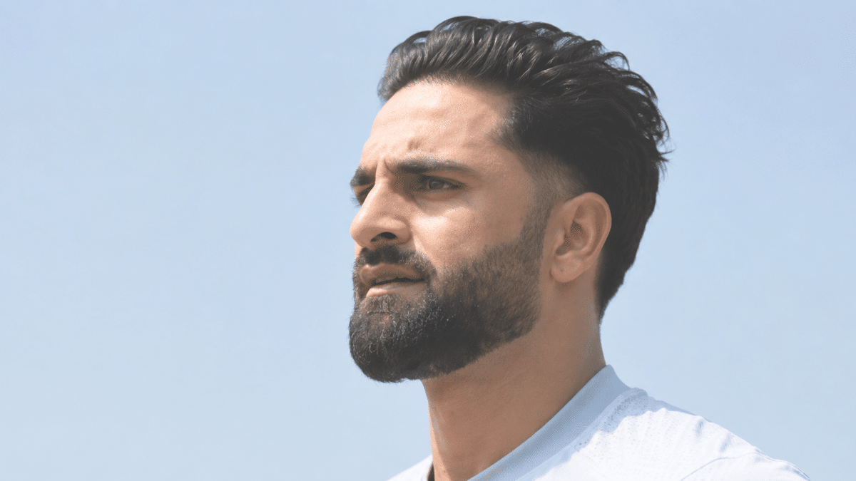 Professional outdoor portrait of a bearded man with styled dark hair wearing a light-colored athletic shirt against a clear blue sky.