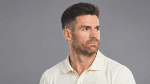Professional three-quarter portrait of English cricketer James Anderson with short dark hair and light stubble, wearing a cream polo shirt against a neutral gray studio background.