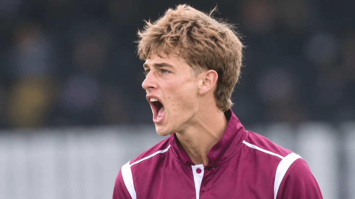 Young male cricketer in a maroon sports jersey, shown in a left-facing three-quarter profile with an intense expression, against a blurred stadium background.