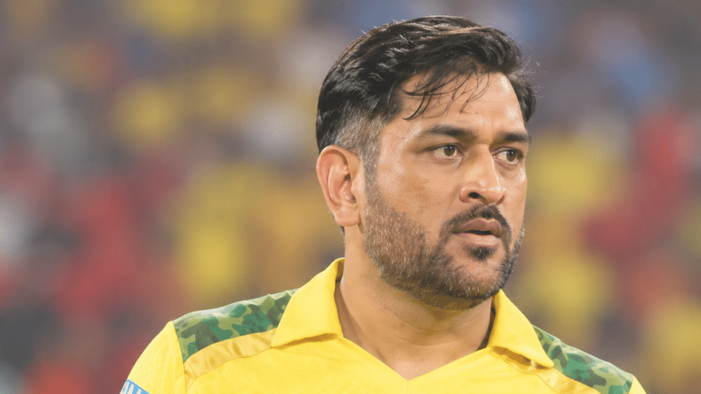 MS Dhoni in a yellow Chennai Super Kings cricket jersey, shown in a right-facing three-quarter profile inside a brightly lit cricket stadium with a blurred crowd in the background.