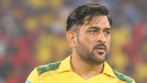MS Dhoni in a yellow Chennai Super Kings cricket jersey, shown in a right-facing three-quarter profile inside a brightly lit cricket stadium with a blurred crowd in the background.