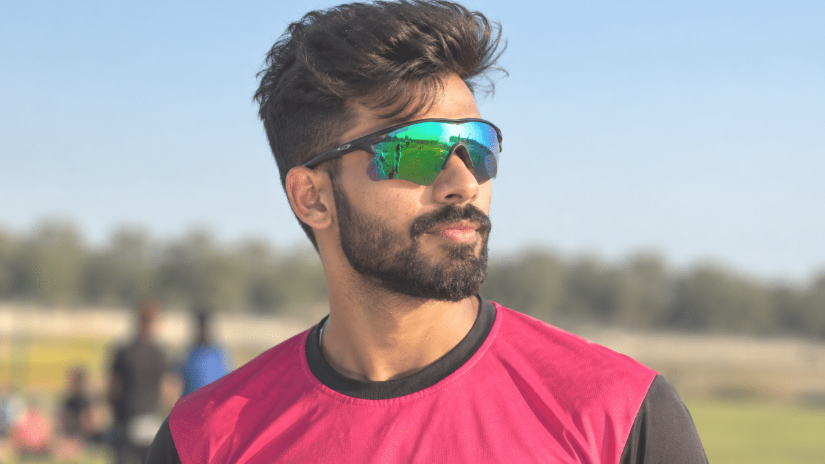 Chinmay Sutar, Indian domestic cricketer, wearing a magenta sports jersey and green mirrored sunglasses, looking to the right with a neatly trimmed beard and styled dark hair against a softly blurred outdoor cricket ground.