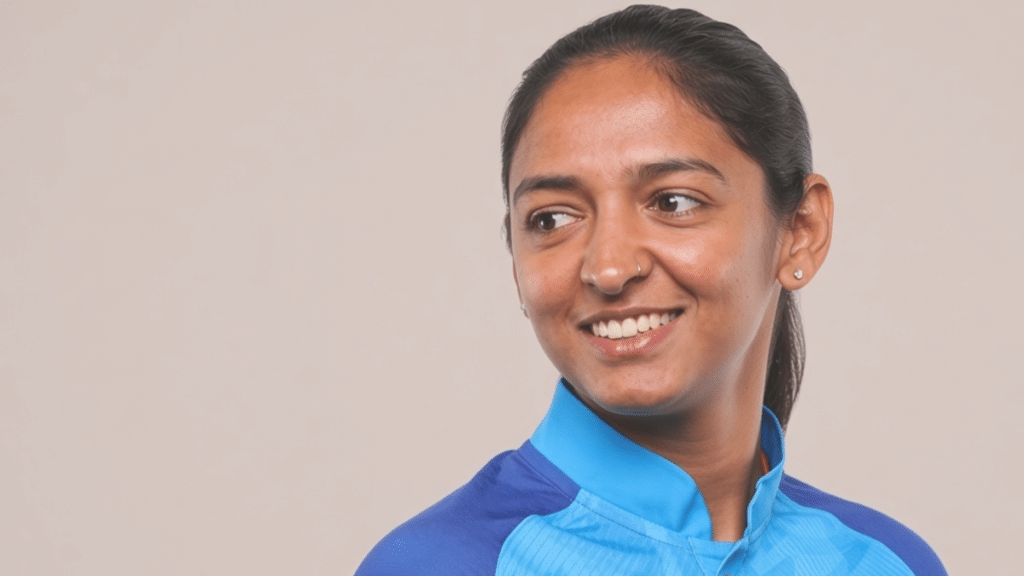 Harmanpreet Kaur smiling and looking to the left, with dark hair pulled back, wearing a blue sports jersey against a plain beige background.