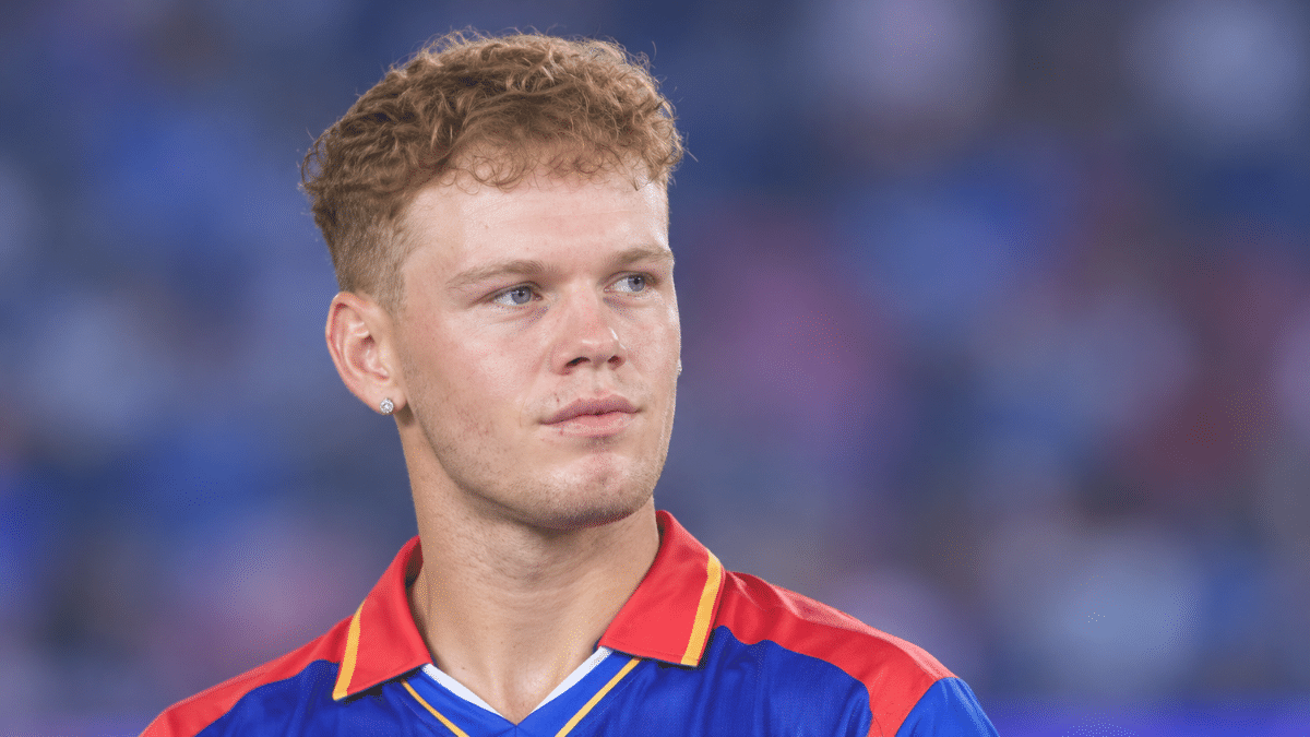 Jake Fraser-McGurk, Australian cricketer, wearing a blue jersey with red and yellow accents, looking to the right against a blurred blue and purple background.