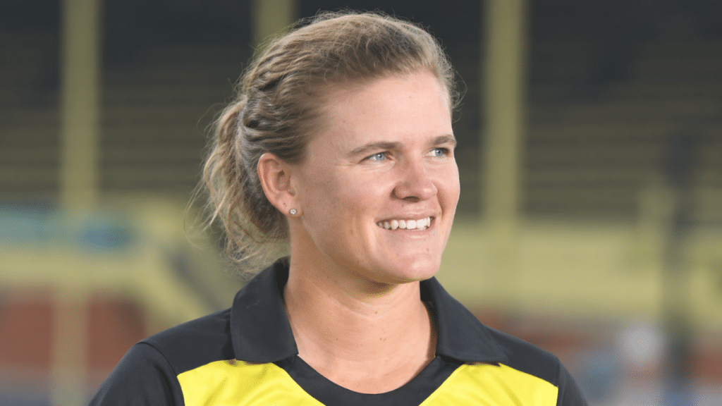 Jess Jonassen, Australian women’s cricketer and left-arm spin-bowling all-rounder, smiling in a black-and-yellow cricket jersey against a blurred stadium background.