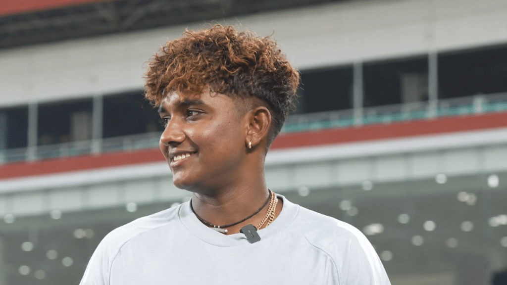 G. Kamalini, Indian women’s cricketer and wicketkeeper-batter, wearing a light grey sweatshirt with curly, brown-highlighted hair and gold jewellery, smiling while looking to the left against a blurred stadium background.