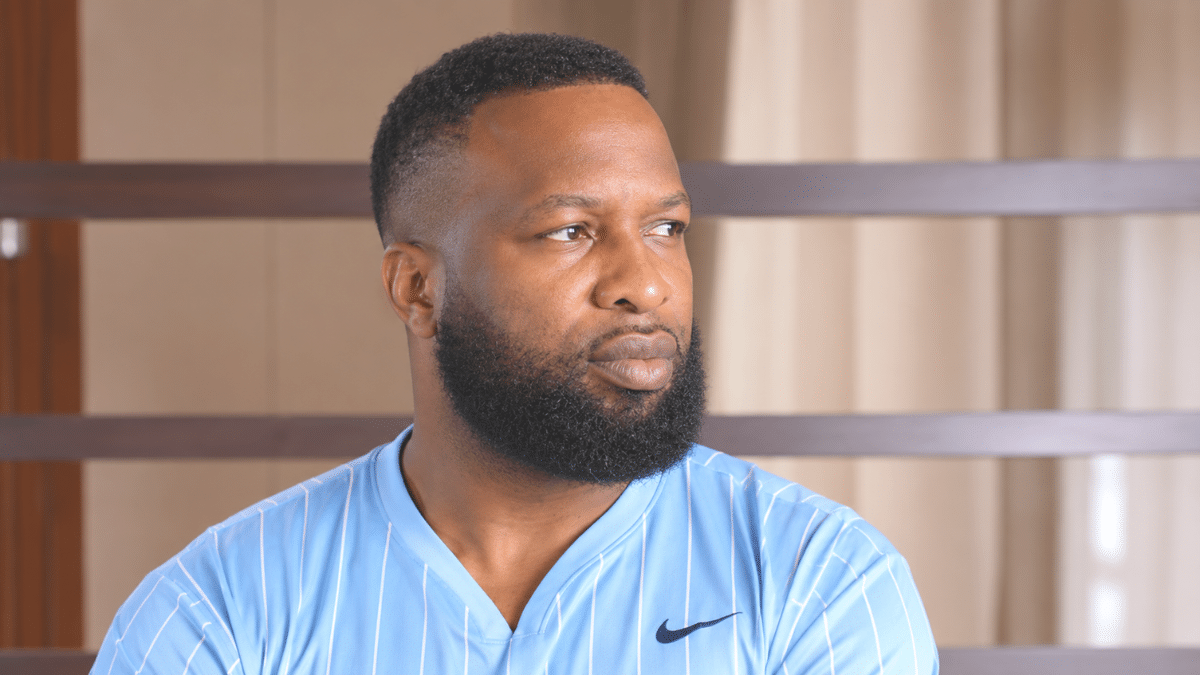 Kieron Pollard, former West Indies cricket captain and powerful all-rounder, wearing a light-blue pinstriped sports jersey with a full black beard, looking to the right against a softly blurred indoor wooden background.