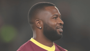 Kieron Pollard looking to the right with a slight smile, short hair and full beard, wearing a maroon jersey with yellow collar against a blurred dark background.