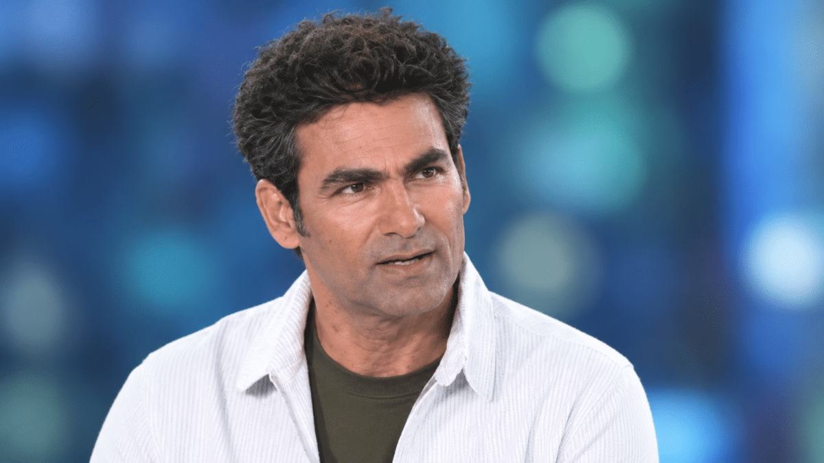Mohammad Kaif, former Indian cricketer, wearing a white pinstriped shirt over a dark olive T-shirt, looking slightly to the right against a blurred blue and teal background.