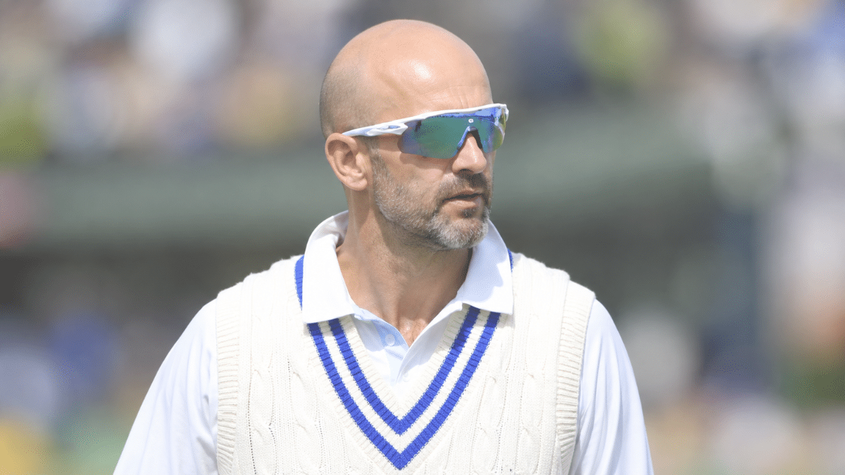 Nathan Lyon, Australian international cricketer and right-arm off-spin bowler, wearing a white cricket shirt and cream sweater vest with blue stripes, looking to the right behind mirrored sunglasses.