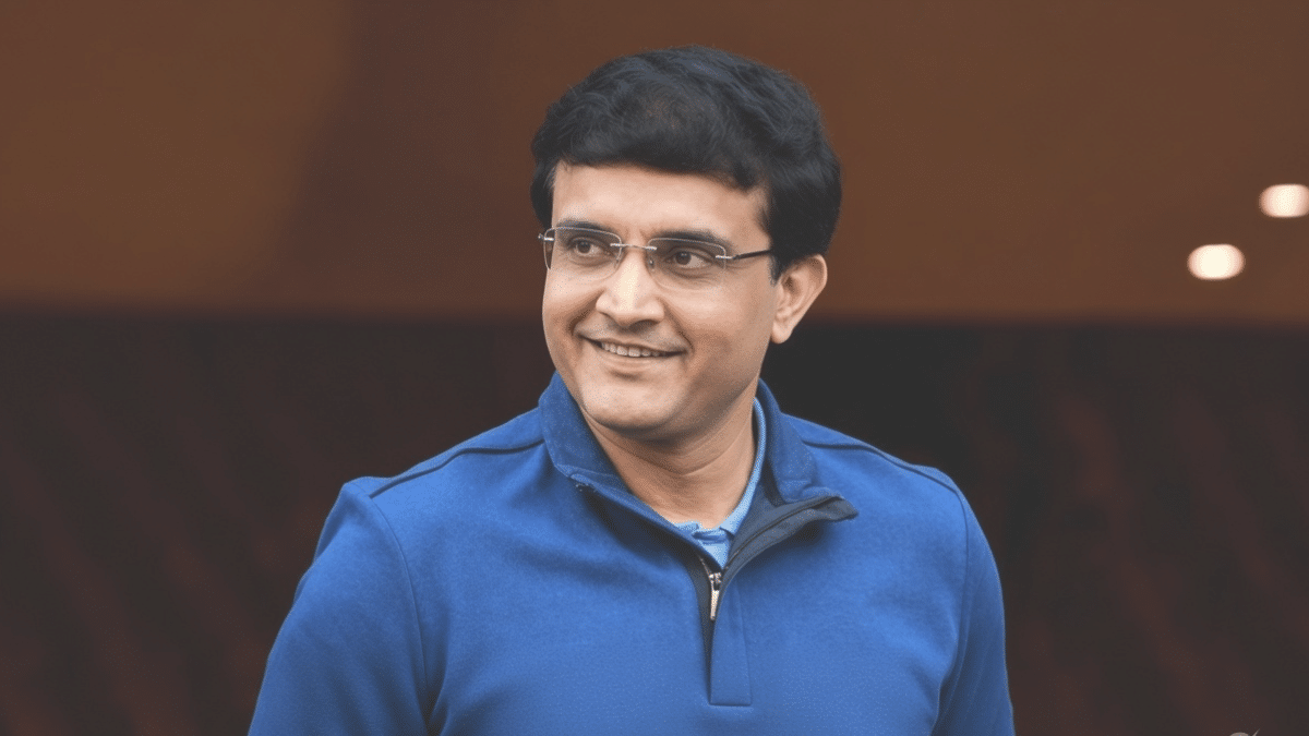 Sourav Ganguly smiling and looking slightly to his left, wearing rimless glasses and a blue quarter-zip sweater against a dark brown background.