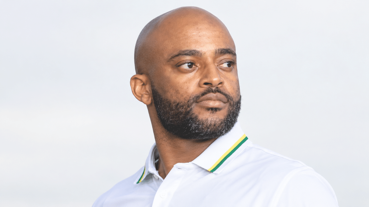 Temba Bavuma, South African international cricketer and captain, wearing a white polo shirt with green-and-yellow collar stripes, looking to the right with a neatly trimmed beard against a clean, light background.