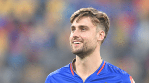 Will Jacks, England cricket all-rounder, smiling in a royal-blue cricket jersey with red and gold trim, looking slightly upward against a blurred stadium background.
