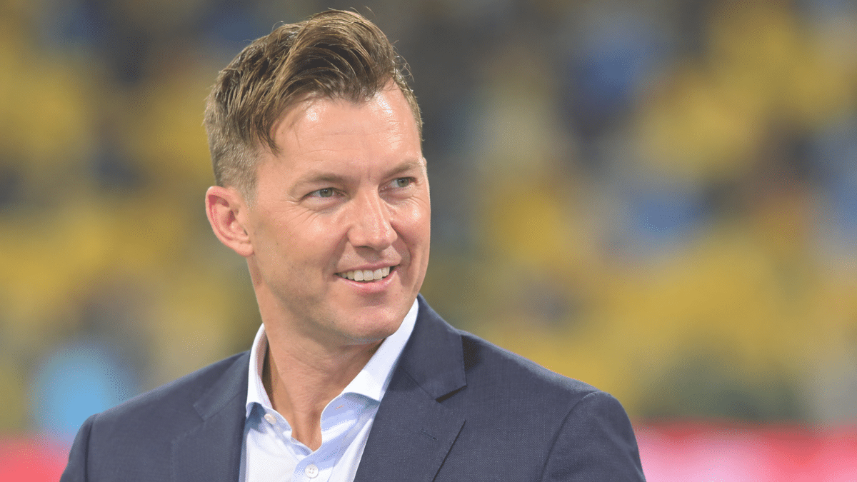 Brett Lee, former Australian fast bowler, wearing a navy blue suit jacket over a light blue shirt, smiling and looking to the right against a blurred yellow stadium background.
