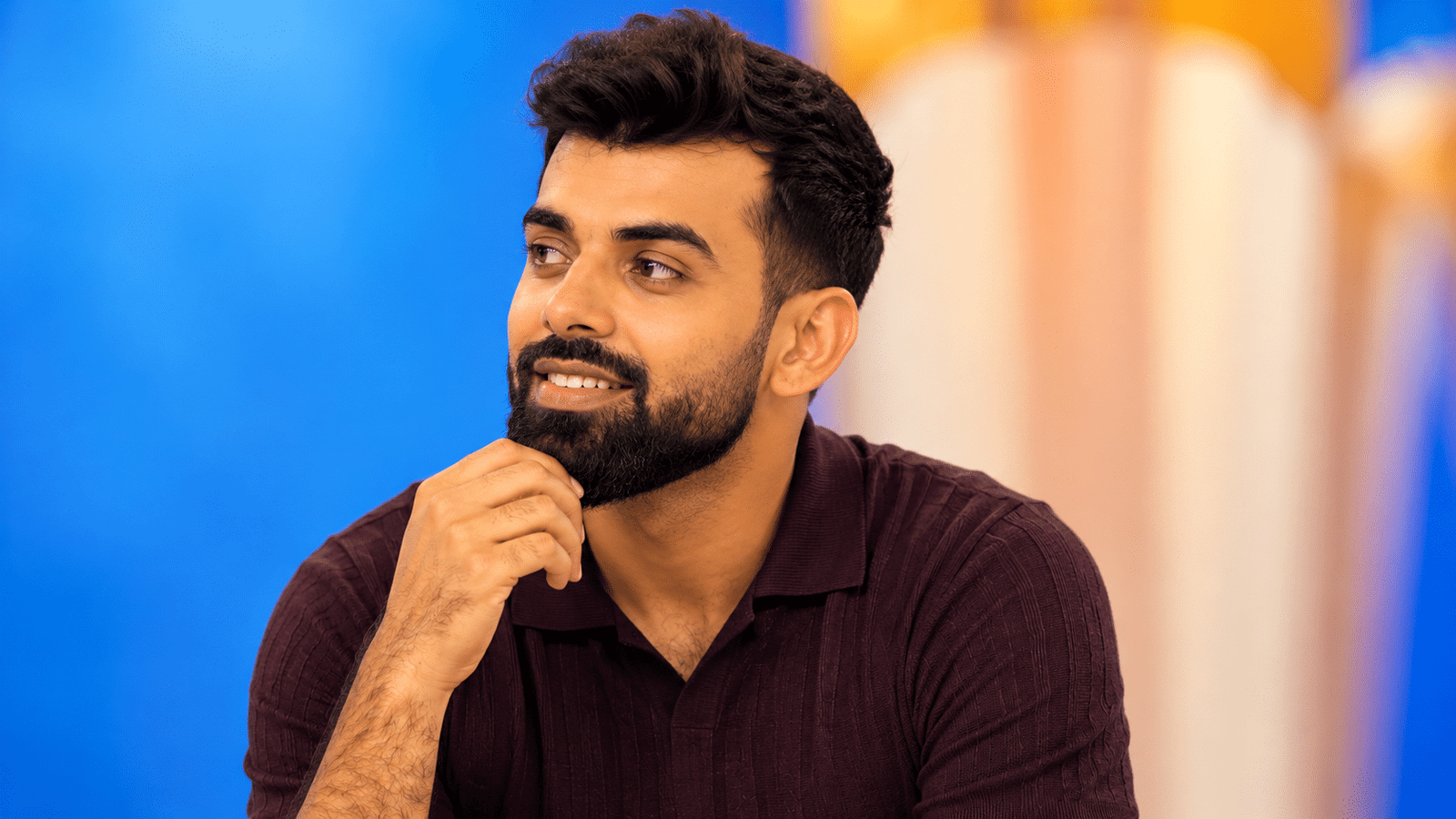 Shadab Khan, Pakistani cricket all-rounder and leg-spin bowler, wearing a burgundy polo shirt and looking to the left with his hand near his chin against a blue and orange background.