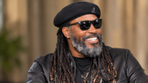 Chris Gayle, former West Indies cricket star known as the “Universe Boss,” smiling and looking to the right while wearing a black beret, dark sunglasses, a black leather jacket, and layered necklaces, with long dreadlocks and a salt-and-pepper beard against a softly blurred background.