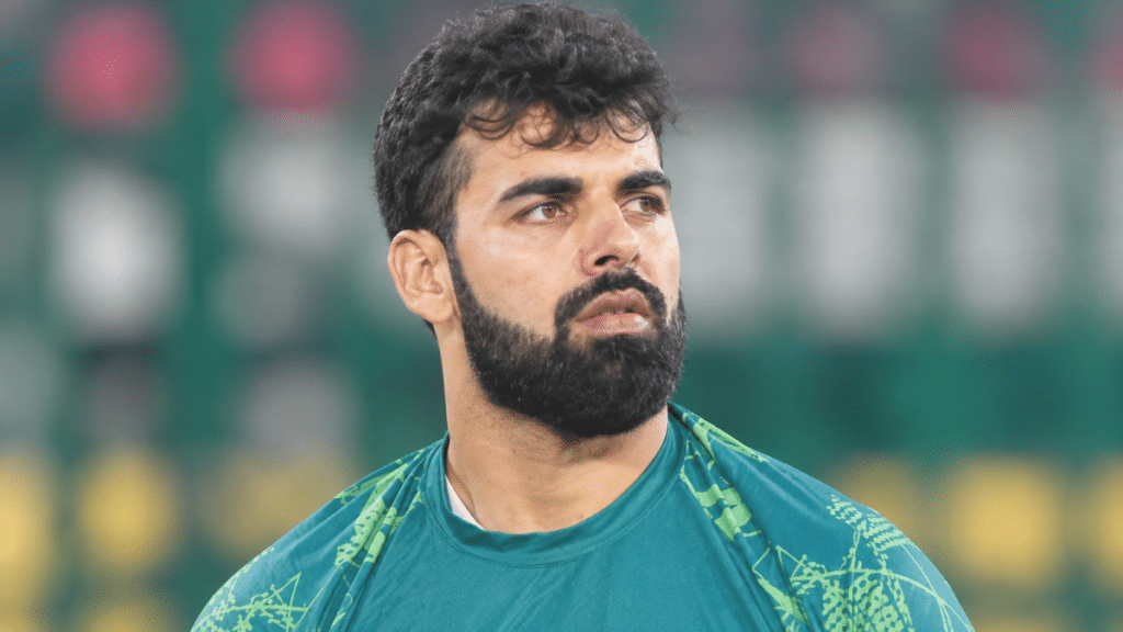 Shadab Khan, Pakistan international cricketer and leg-spin bowling all-rounder, wearing a teal-green cricket jersey with lime-green patterns, looking to the right against a blurred cricket stadium background.