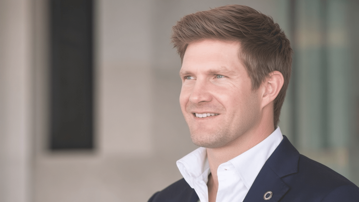 Shane Watson smiling and looking to the left, with short light brown hair, wearing a white shirt and navy blazer against a blurred indoor background.