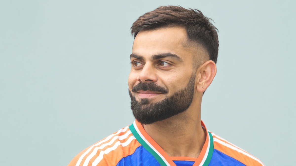 Virat Kohli smiling in a blue and orange jersey against a plain background.